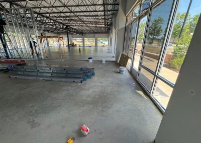The large room, a vision of Nextt Commercial Builders, is under construction with metal frames, ladders, and tools scattered about. Large windows reveal trees outside as the transformation journey unfolds in this commercial construction phase.